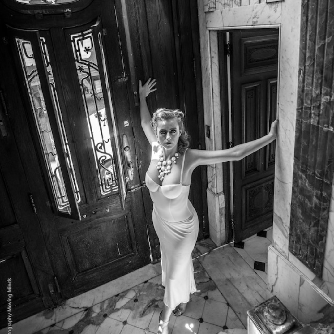 Sara Rose, the tango dancer, is looking up at the camera, her hands on two door frames, stretched out behind her. She is wearing a white dress.